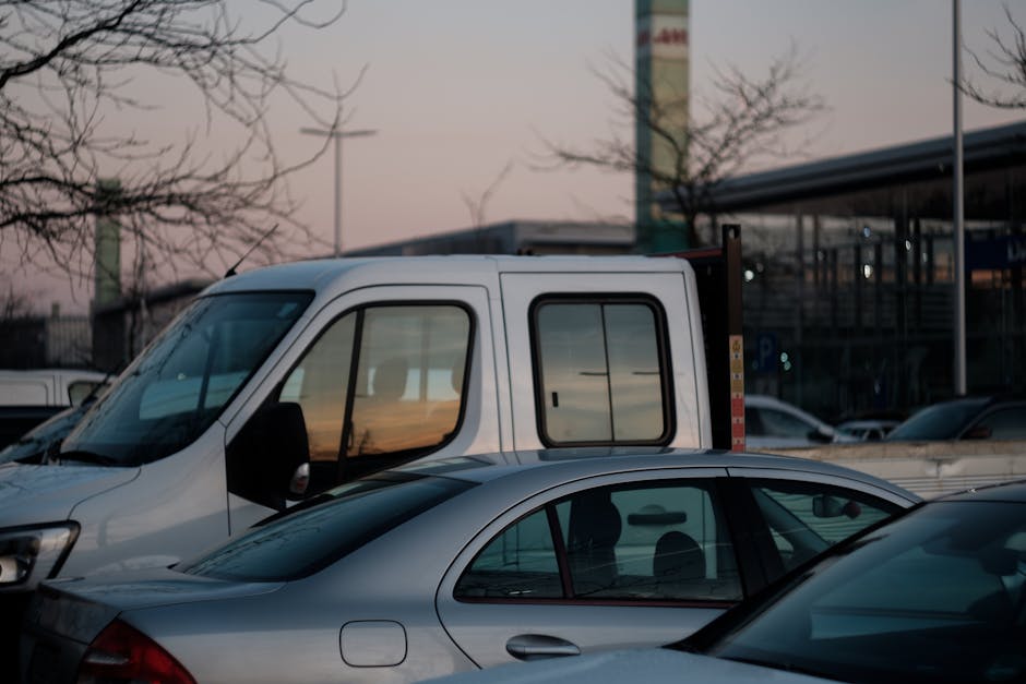A white flatbed van with a lowered tailgate is parked among other vehicles in a parking lot during dusk, with a clear sky and soft sunset hues visible in the background. The van is positioned near a modern building with large glass windows and a covered area, possibly a retail or commercial space. Inside the van, partially visible, are packing materials such as cardboard boxes, plastic wrapping, and blankets, indicating a home relocation or furniture transport process. Adjacent to the van, a silver sedan and a grey car are also parked, with the grey car’s rear window reflecting the surrounding environment. In front of the white van, a black trolley may be visible, suggesting ongoing loading or unloading activities. The scene includes leafless trees with bare branches, highlighting an autumn or winter season. The presence of the Man with Van Hampton Wick logo and the context of house removals and logistics is implied by the setting, supporting the topic of packing and moving services essential for complying with parking and permit rules during a household move.