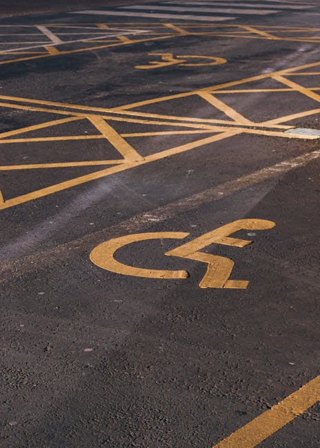 A close-up view of a paved parking lot featuring multiple designated space markings painted in yellow, including several with the wheelchair symbol indicating accessible parking. The surface shows signs of wear and slight discoloration, with some areas appearing more textured than others. In the background, additional parking spaces are visible, with clearly marked lines and symbols, alongside a van partially visible on the right side of the image, suggesting a moving or transport context associated with house removals. This scene illustrates the importance of understanding parking regulations for safe and compliant furniture transport during home relocation processes, as managed by a professional removal service like Man with Van Hampton Wick.