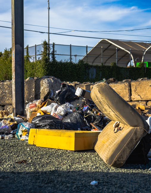 An outdoor scene showing a pile of bulky waste, including a large, worn-out beige sofa with visible fabric texture and signs of use, positioned at an angle with one arm rest leaning against the ground. Surrounding the sofa are several black plastic garbage bags filled with household waste, some torn open revealing crumpled paper and plastics. A yellow plastic storage container lies nearby, partially covered by the waste. Behind the waste, a low stone wall separates the area from a fenced property with a white curved greenhouse structure and some greenery. A wooden utility pole with overhead power lines is situated on the left side of the image, under a bright blue sky with scattered clouds, indicating daylight. The scene captures waste collection or disposal activities relevant to home relocation or furniture transport, as managed by companies like Man with Van Hampton Wick, with the waste awaiting removal from a property to a local waste facility.