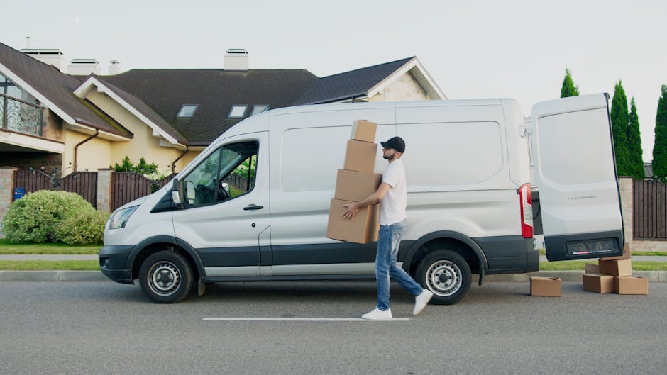 A vintage white small van, used for home relocation, is moving along a roadway with a blurred background that suggests motion. The van's cargo area is loaded with various household furniture pieces, i
