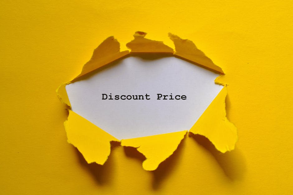 A piece of bright yellow paper is torn open in the center, revealing a white sheet underneath with the black text 'Discount Price' printed on it. The torn edges of the yellow paper are jagged and curling outward, creating a rough circular opening. The background is also yellow, matching the torn paper, and the scene is illuminated evenly, highlighting the texture of the torn paper and the contrasting colors. This image visually conveys the concept of revealing or discovering a reduced cost, relevant to house removals and moving services offered by Man with Van Hampton Wick, as it relates to cost considerations and quotes for relocation projects.