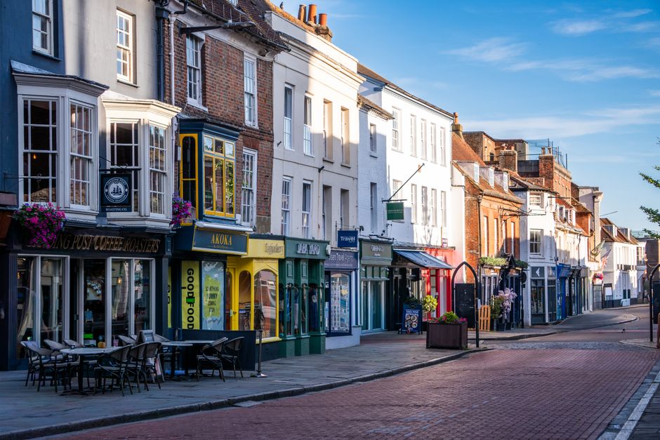 Two men are engaged in loading a moving truck parked on the street outside a shop titled 'Guang' in Hampton Wick. One individual is holding a plastic crate filled with various items, while the other i