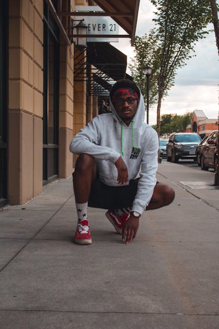 A person crouching on a city sidewalk near a building advertises the Ever 21 store. They are wearing a light grey hoodie with a green drawstring, black shorts, red sneakers, white socks with red and black patterns, and red-tinted sunglasses. Behind them, parked cars are visible along the street, with trees and additional buildings further in the background. The pavement is concrete, and the building frontage includes a window grille and an overhead sign. This scene suggests an urban environment where house removals or moving services, such as those provided by Man with Van Hampton Wick, may involve logistical planning for transporting furniture and boxes, although no furniture or moving equipment is directly visible in the image.