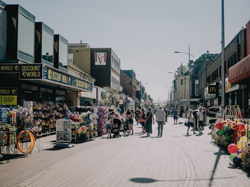 A wide view of Hampton Wick High Street during daytime shows a pedestrianised street lined with local shops, including a discount store and a McDonald's. The pavement is populated with people walking, some pushing strollers, carrying shopping bags, or browsing the street stalls. The stalls on both sides display various goods, such as colorful balloons, toys, and flowers, with items arranged on tables, racks, and carts. Storefronts have signage, with some featuring awnings, and a row of street lamps lines the street. The scene captures a typical busy shopping day, with natural daylight illuminating the area. Durings home relocation or furniture transport, such streets often serve as the pathway for delivery vehicles or removals vans, which can be parked nearby while items are being carried to or from properties. This image, taken through the lens of a professional removals company like Man with Van Hampton Wick, reflects the bustling environment where packing, loading, and transporting household goods occurs in close proximity to local shops and community spaces.
