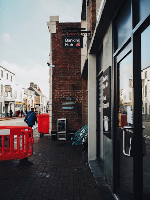 A person crouching on a city sidewalk near a building advertises the Ever 21 store. They are wearing a light grey hoodie with a green drawstring, black shorts, red sneakers, white socks with red and black patterns, and red-tinted sunglasses. Behind them, parked cars are visible along the street, with trees and additional buildings further in the background. The pavement is concrete, and the building frontage includes a window grille and an overhead sign. This scene suggests an urban environment where house removals or moving services, such as those provided by Man with Van Hampton Wick, may involve logistical planning for transporting furniture and boxes, although no furniture or moving equipment is directly visible in the image.