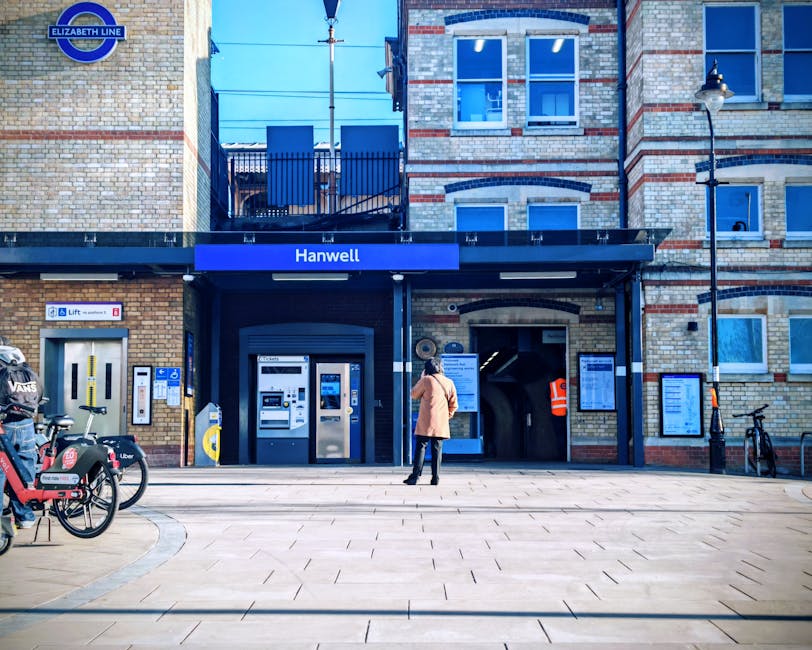 A woman stands outside the Hanwell station entrance, part of the Elizabeth Line, on a clear day with bright blue sky and sunlight illuminating the brick facade. The station sign is prominently displayed above the entrance, which features a dark canopy and a roller shutter door. To the left, there are several bicycles parked, including one with a cargo rack and company branding. Adjacent to the station, there are ticket machines, a yellow contactless payment device, and signage with directional information. On the right side, a black street lamp and a bicycle stand with an upright bike are visible. The scene depicts urban infrastructure with a clean, tiled pedestrian area, suitable for walking and loading transport items. The image relates to the logistics of home relocation or moving services, such as those offered by Man with Van Hampton Wick, highlighting a typical urban transit point used during furniture transport or packing and moving activities connected to house removals around the Hampton Wick area.