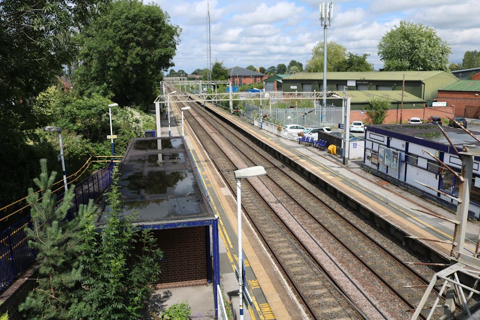 An elevated view of Hampton Wick train station showing multiple railway tracks running parallel across the platform area, with a station building on the right featuring signage and information boards. The platform is partially visible with a few parked cars and a small car park area adjacent to the station. In the foreground, there is a blue metal railing fencing the platform, alongside a small shelter with a flat roof that appears wet, possibly from rain, with reflections visible on its surface. Tall utility poles with overhead wires run along the tracks, and surrounding trees and greenery are visible on the left side of the image, creating a natural backdrop. The scene depicts a typical urban railway environment during daytime with partly cloudy skies, relevant to a house relocation context involving furniture transport and moving logistics delivered by services such as Man with Van Hampton Wick.