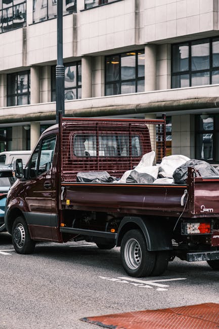A vintage beige small delivery van is in motion on a road during daytime, with its exterior loaded with various household items prepared for a home relocation. The vehicle's bed is filled with stacked