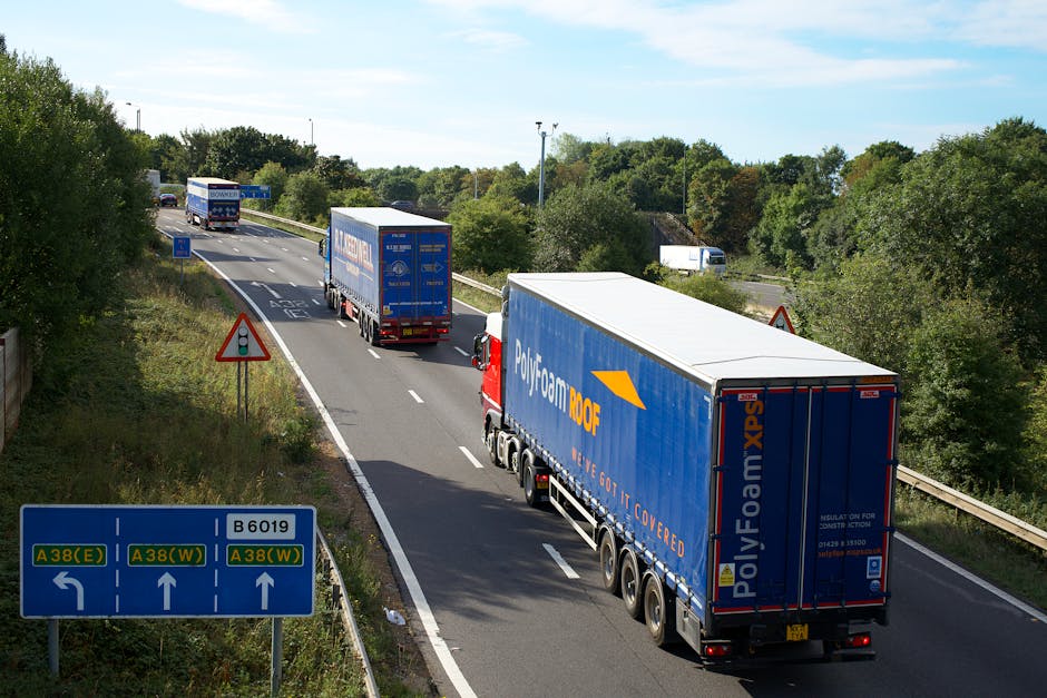 A line of three large articulated lorries with blue Polyfoam branding on their trailers are traveling along a dual carriageway bordered by lush green trees and bushes. The closest truck is positioned on the right side of the image, with a white cab and multiple black wheels, and appears to be in the process of transporting household or commercial goods as part of a removal or relocation service. The second truck is further ahead, also with a white cab and a similar blue trailer, followed by a third truck, all aligned in the same direction. The road features white dashed lane markings and a red and white triangular roadwork warning sign on the side, indicating possible construction or caution. In the foreground, a green roadside sign displays directions to the A38 (E and W) and B6019, situated near the edge of the verge, which is covered with grass and low shrubs. The background includes more trees and a partly cloudy sky, suggesting fair weather conditions suitable for a home relocation or furniture transport operation recognized by Man with Van Hampton Wick, who may be involved in such logistics.