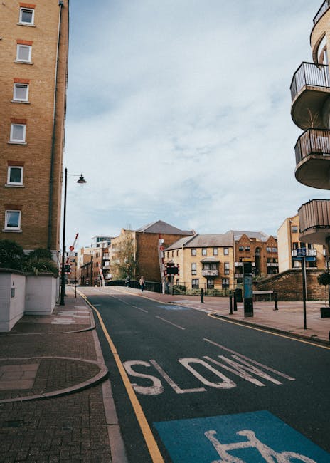 A narrow residential street in Hampton Wick during daytime, with multi-storey brick buildings on both sides, some featuring balconies with metal railings. The street is paved with asphalt and has a yellow line marking the edge adjacent to the sidewalk. A prominent white 'STOP' marking is painted on the road near the foreground, with a blue parking space marking nearby. Streetlights and bollards line the sidewalk, and a few pedestrians can be seen in the distance. The sky is cloudy with patches of blue. This scene is representative of the urban environment where house removals, such as those performed by Man with Van Hampton Wick, often take place, especially involving home relocation or furniture transport through tight city streets.