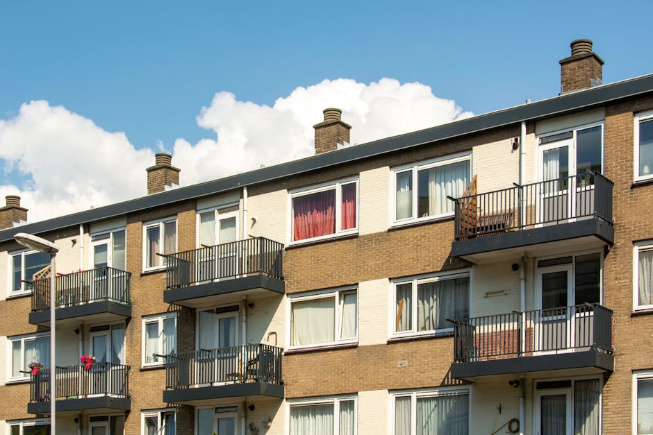 A multi-storey residential building with three floors, featuring a brown brick facade and white exterior panels surrounding the windows. Each floor has several glass windows, some with curtains, and small balconies with black metal railings. The balconies contain outdoor furniture, including chairs, and potted plants or decorations. The building's roof has four brick chimneys, and a streetlamp is partially visible at the left edge of the image. The sky above is partly cloudy with a bright blue backdrop. The scene captures a typical residential flats exterior, relevant for home relocation or moving services, with clear visual details of the building's structure and balcony layout, supporting the context of house removals and furniture transport around flats without lifts, as indicated on the page about solutions for such moves. The image has natural lighting and no people are visible in the scene.