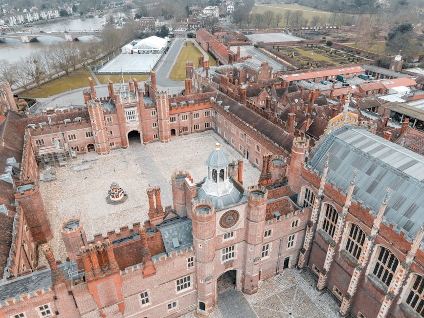 An aerial view of a historic red brick castle with multiple turrets and a large central courtyard, surrounded by a gravel pathway. The castle features detailed stonework, tall chimneys, and large windows, with some sections still under scaffolding. In the background, there are landscaped gardens, a sports field, and a river with a bridge crossing over it. The scene includes a moving van parked near the castle with a black exterior and a visible rear door, alongside a few cardboard boxes and wrapped furniture items placed inside the van, indicating a home relocation or furniture transport process. The overall environment is outdoor, under natural daylight, with an expansive view that captures both the castle and its surrounding landscape. This image exemplifies the logistics involved in house removals, as handled by companies like Man with Van Hampton Wick, with a focus on packing, loading, and transport in a historic residential setting.