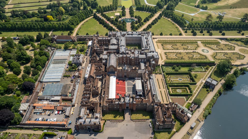 An aerial view of Hampton Court Palace showing its historic brick and stone facade, with the main entrance gate in the foreground. The palace is surrounded by well-maintained formal gardens featuring symmetrical flower beds, trimmed hedges, and gravel pathways. To the left of the palace, there is a large loading area with several parked vehicles including a white van, and a variety of cardboard boxes, wrapped furniture, and packing materials being prepared for relocation. A team of movers from Man with Van Hampton Wick is visible, carefully lifting and arranging furniture such as chairs and small cabinets onto the van for transport. The scene is set during daytime with clear natural lighting, highlighting the detailed brickwork and lush greenery of the gardens, reflecting the process of home relocation and the logistical planning involved in a furniture transport or moving service near Hampton Court Palace.
