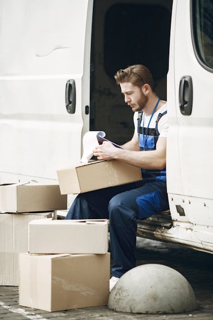 A man wearing a striped T-shirt and blue work overalls is sitting inside the open door of a white van parked on a paved area. He is looking at a clipboard or sheet of paper while holding a cardboard b