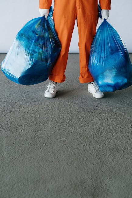 A person wearing orange overalls and white sneakers is standing indoors on a grey carpeted floor, holding two large blue plastic garbage bags filled with waste materials. The person's hands, covered with gloves, grip the bags at each side. In the background, a plain light grey wall is visible, and the individual appears to be in the process of loading the bags for disposal or transport, which could relate to home relocation or packing activities. The image highlights the manual handling of waste during a house removal or moving service, illustrating proper material collection and logistics involved in disposal steps. A company named Man with Van Hampton Wick, specialising in removals, is implied to be involved in managing such tasks as part of their comprehensive relocation services.